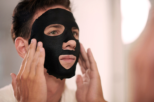 Face of young man applying charcoal sheet mask