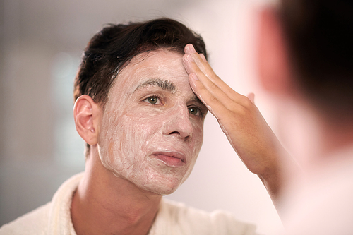 Young man with dark hair applying rejuvenating face mask in front of mirror in bathroom after washing with gel or foam in the morning