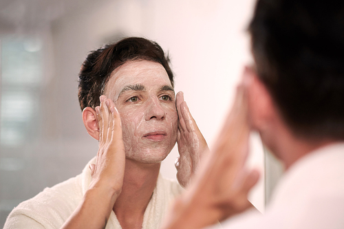 Young man washing face with cleansing foam in the evening