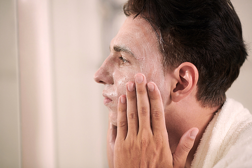 Side view of young man applying purifying or rejuvenating mask on his face while standing in front of mirror and taking care of himself