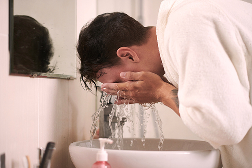 Young man in soft white bathrobe bending over sink in front of mirror in bathroom and washing his face during daily hygiene procedures