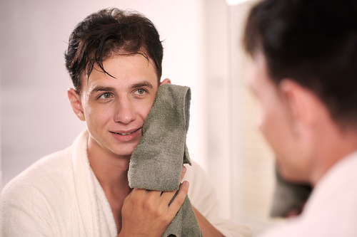 Young smiling man in white soft bathrobe drying his face with towel after washing and looking at his reflection in mirror in the bathroom