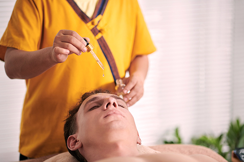 Hand of young female cosmetician dropping essential oil or serum on face of relaxed male client lying on couch during skincare procedure