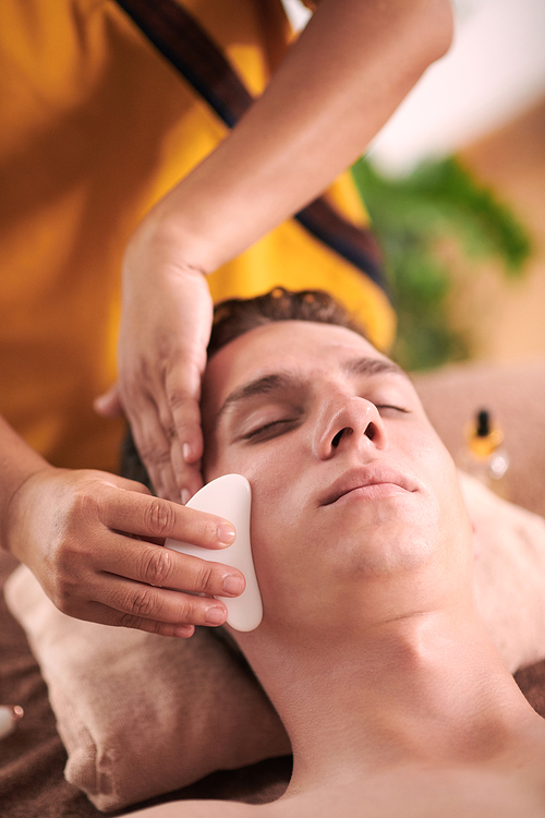 Young man getting gua sha face massage to relieve tension in face and reduce puffiness