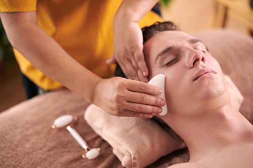 Young female beautician with guasha smoothing skin of male client face during luxurious spa procedure while standing behind his head