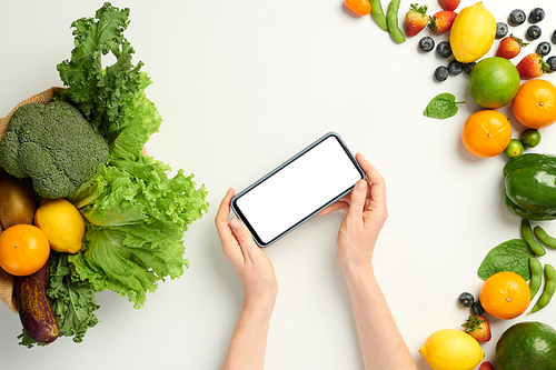 Hands of person holding smartphone with white screen over table with fresh organic groceries from supermarket