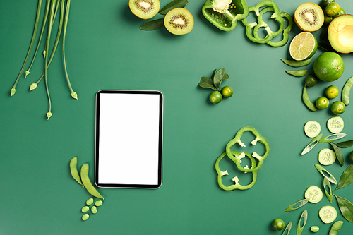 Tablet computer with empty screen on table with green fruits and vegetables prepared for cooking