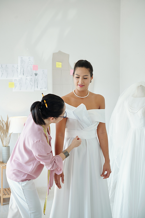 Seamstress checking wedding dress on bride during fitting in atelier