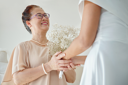 Happy senior woman touching hands of daughter standing in wedding dress ready for ceremony