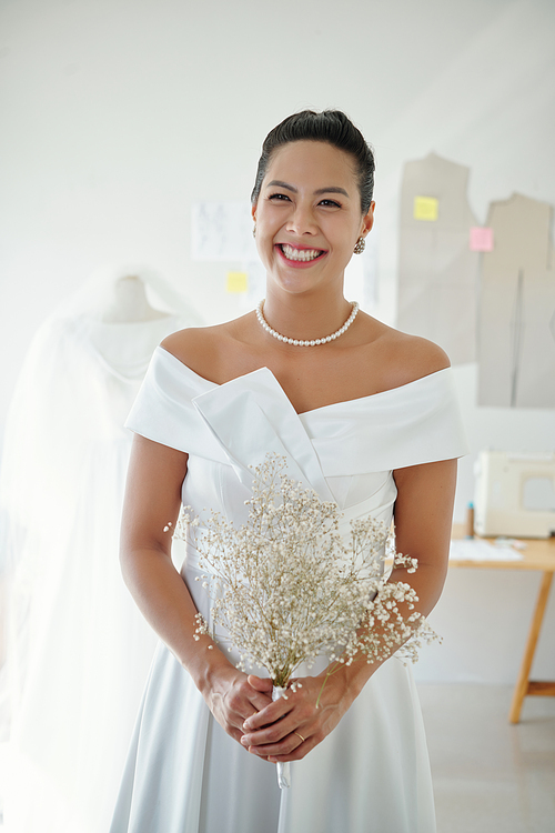 Portrait of happy bride with flowers in hands standing in fashion atelier