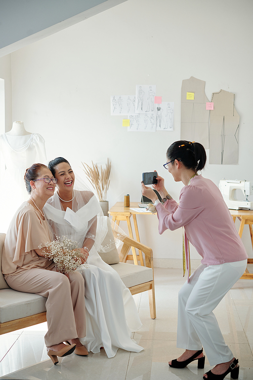 Joyful bride and her mother asking tailor to take photo after fitting dress