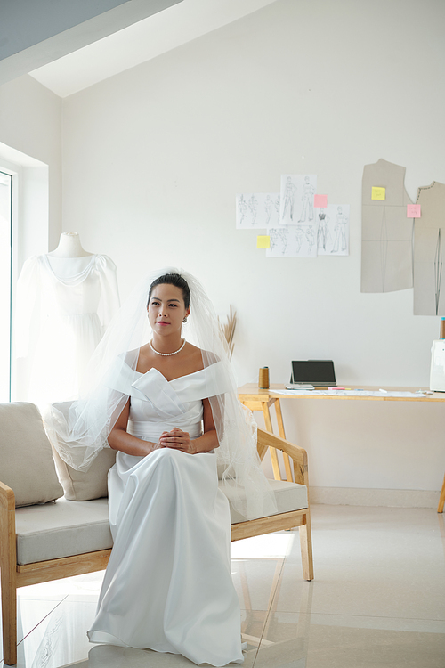 Pensive bride in beautiful wedding dress and veil sitting on fashion atelier