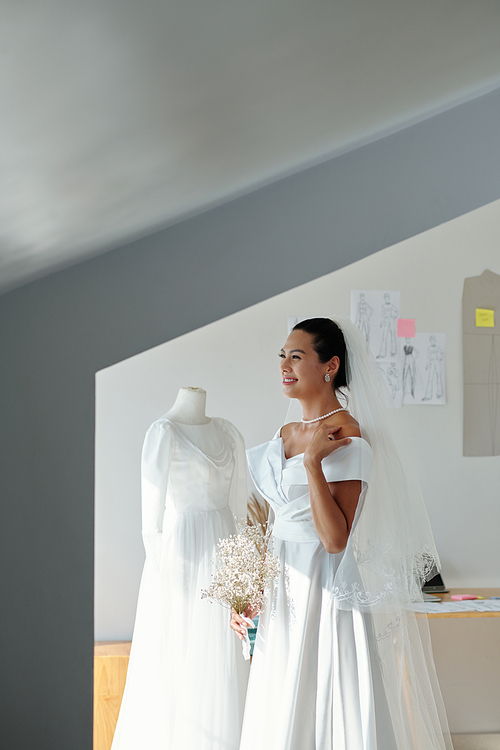 Joyful beautiful bride in white wedding dress standing in salon with bouquet of flowers