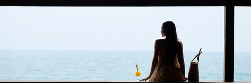 Young woman in flowing dress enjoying beautiful seascape and drinking fruit cocktail