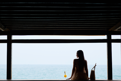 Young woman sitting next to glass of fruit juice and enjoying beautiful seascape