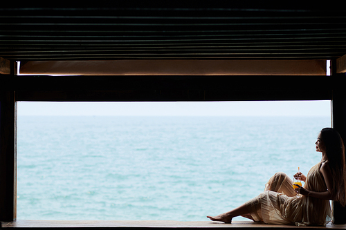 Young woman in silk dress sipping cocktail and looking at beautiful sea