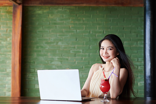 Smiling young woman drinking fruit cocktail when working on laptop at bar counter