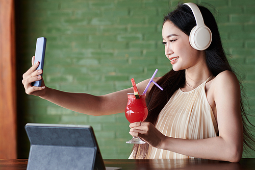 Pretty young woman in headphones taking selfie with fruit cocktail in cafe