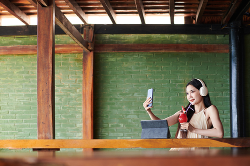 Smiling blogger in headphone taking selfie with fruit cocktail