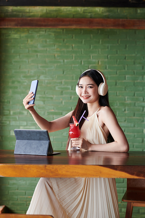 Portrait of smiling young woman having fruit cocktail when video calling friend to discuss news