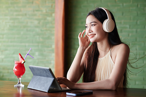 Cheerful young woman in headphones video calling friend when resting in bar