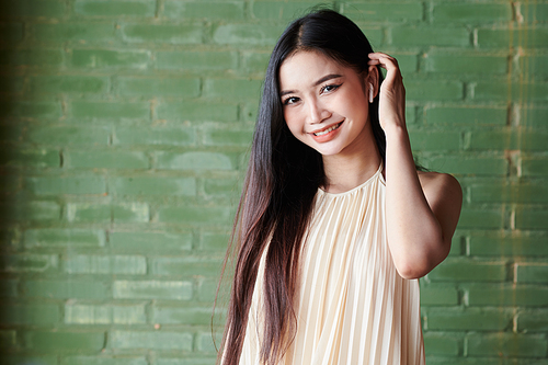 Portrait of lovely young Vietnamese woman tucking hair behind ear and smiling at camera