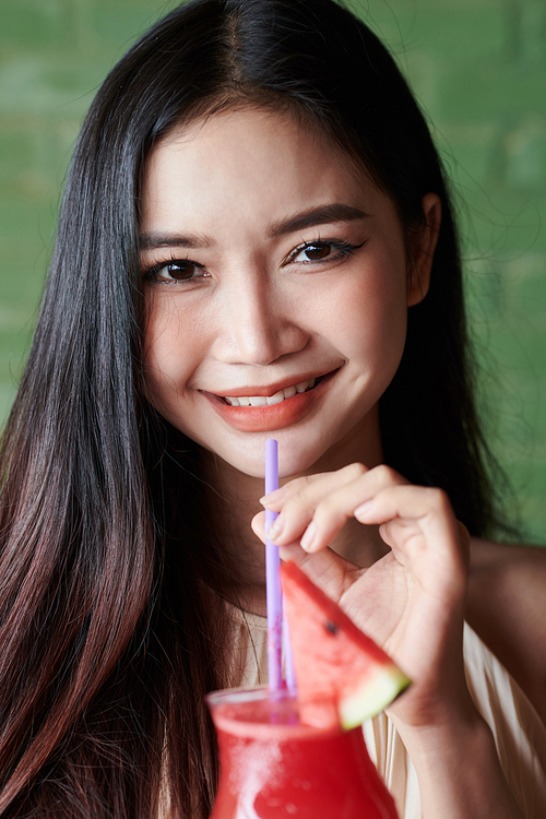 Portrait of joyful young woman drinking watermelon smoothie and smiling at camera