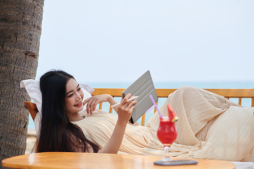 Happy young woman relaxing on patio sofa and watching entertaining videos on tablet computer