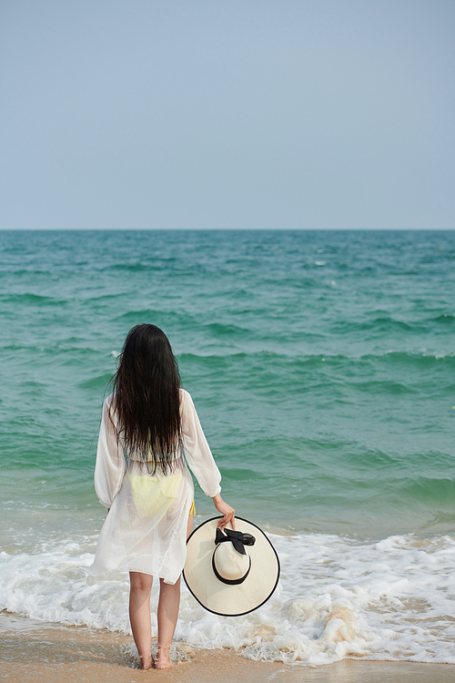 Young woman standing on sandy beach, taking off hat and enjoying sea waves