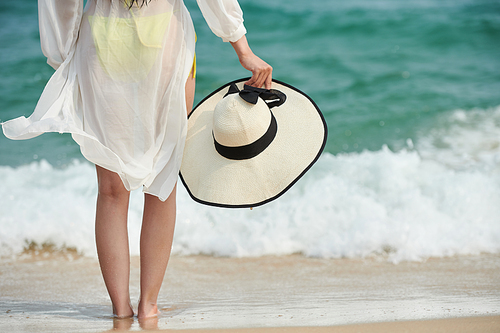 Barefoot young woman standing on sandy beach and enjoying sea waves