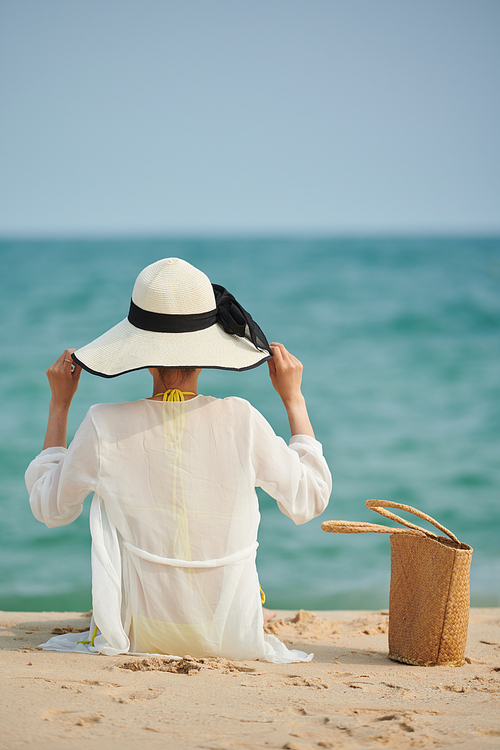 Young woman in straw hat sitting on sandy beach, view from back