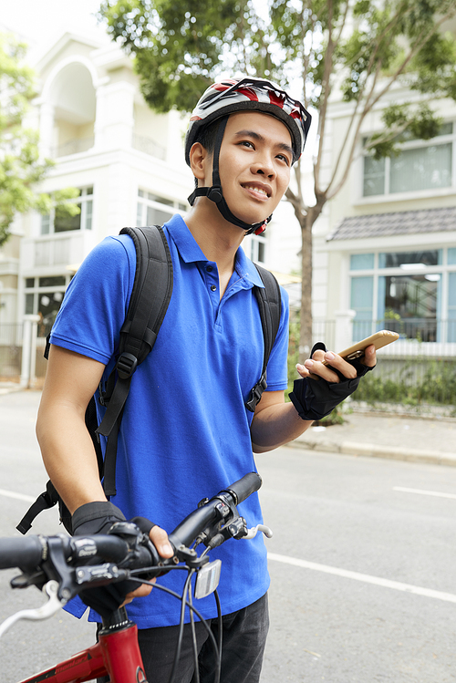 Smiling young Asian courier with smartphone in hand searching for delivery address