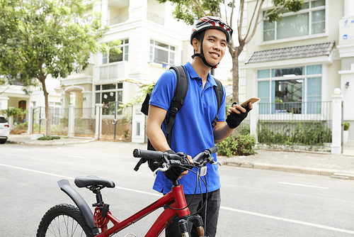 Smiling young Vietnamese delivery man standing next to his bicycle with smartphone in hand