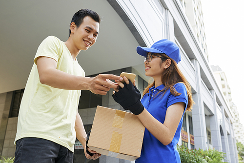 Smiling young delivery woman giving package and asking customer to sign document on her smartphone