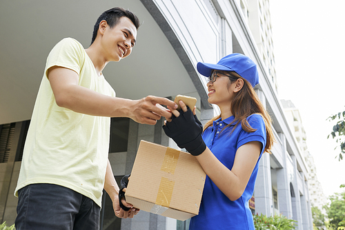 Happy young Vietnamese man receiving package and and signing via mobile application on smartphone in hand of delivery woman