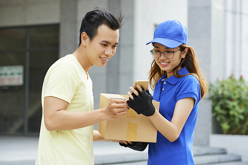 Pretty young delivery woman giving package and asking customer to sign online document on her smartphone