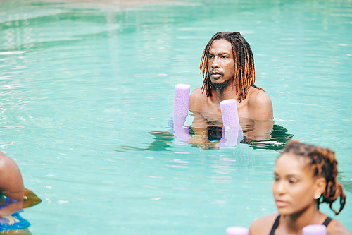 Serious young Black man with dreadlocks exerising in swimming pool with foam noodle in hands