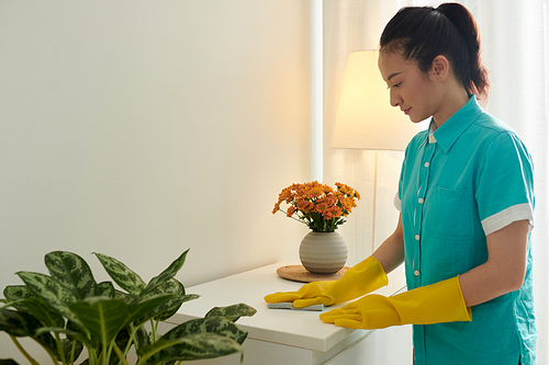 Cleaner girl in uniform and rubber gloves wiping dust from the table during her work in apartment