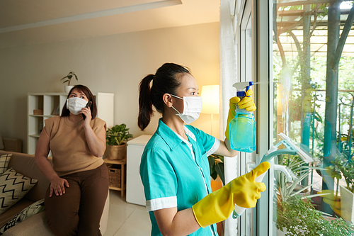 Asian young woman in mask and uniform using spray to clean the windows during her work in apartment