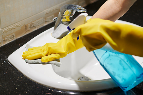 Close-up of woman in protective rubber gloves washing sink with rag and spray in bathroom