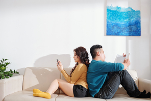 Boyfriend and girlfriend sitting on sofa back to back, texting and reading book