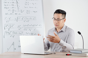 University teacher sitting at desk with opened laptop and recording online class for students