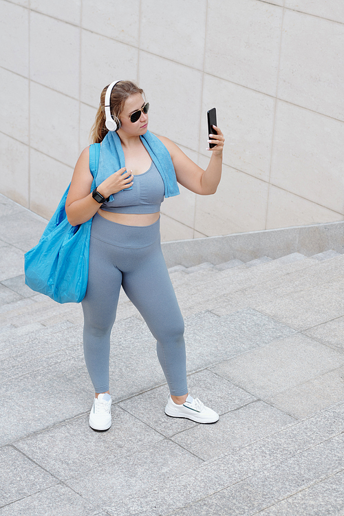 Pretty fit plus size young woman with gym bag posing for selfie after training in sunglasses and headphones