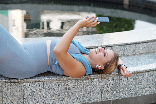 Beautiful smiling fit plus size young woman with smartphone relaxing after outdoor training and checking her social media account