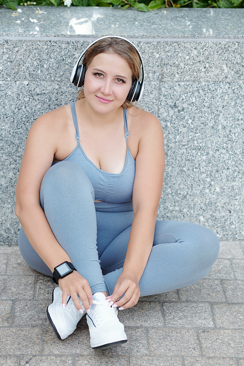 Beautiful smiling plus size young woman in headphones tying shoe laces before morning run