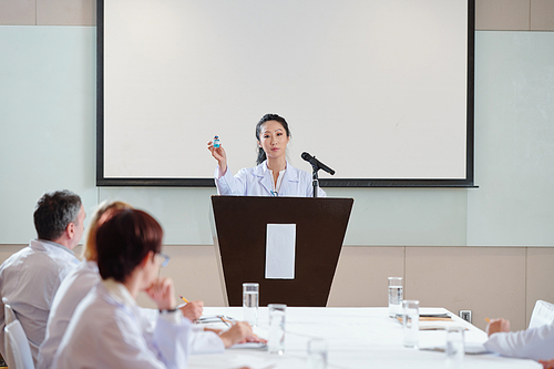 Female young Asian scientist standing at tribune at medical conference and talking about new covid-19 vaccine