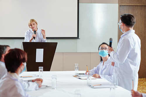 Serious female speaker with vial in hand listening to question from the audience when standing at tribune at medical conference