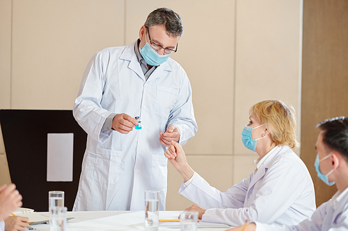 Senior researcher in medical mask showing vial with new vaccine to his colleagues at medical conference