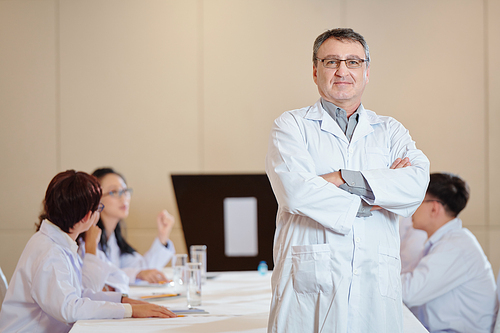 Portrait of smiling confident experienced researcher standing with arms folded, his colleagues discussing work in background