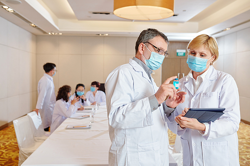 Mature experienced researcher in medical mask showing vial with newest version of vaccine to his coworker when their collegues having discussion i background